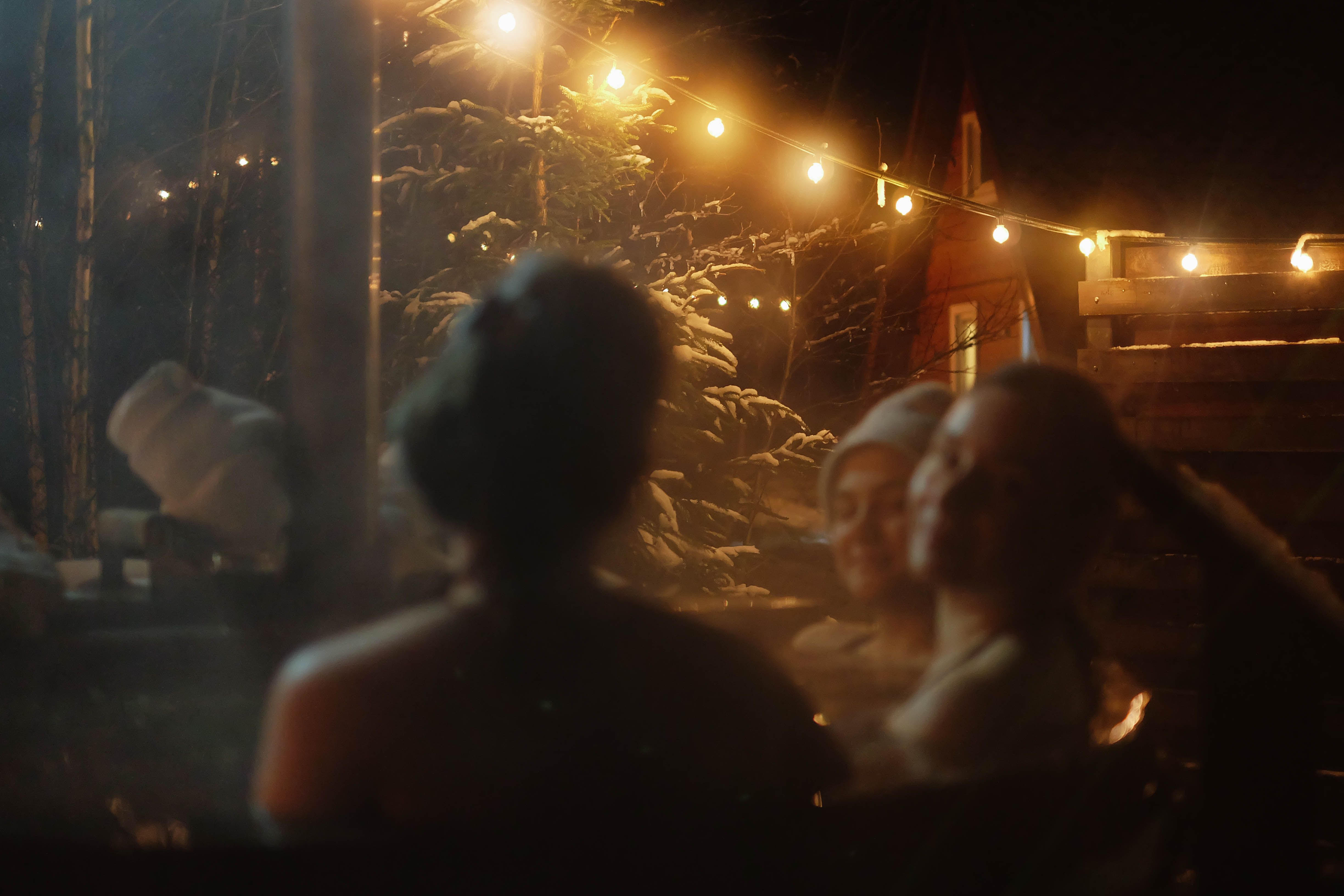 People relaxing in an outdoor hot tub at night surrounded by snow and warm string lights